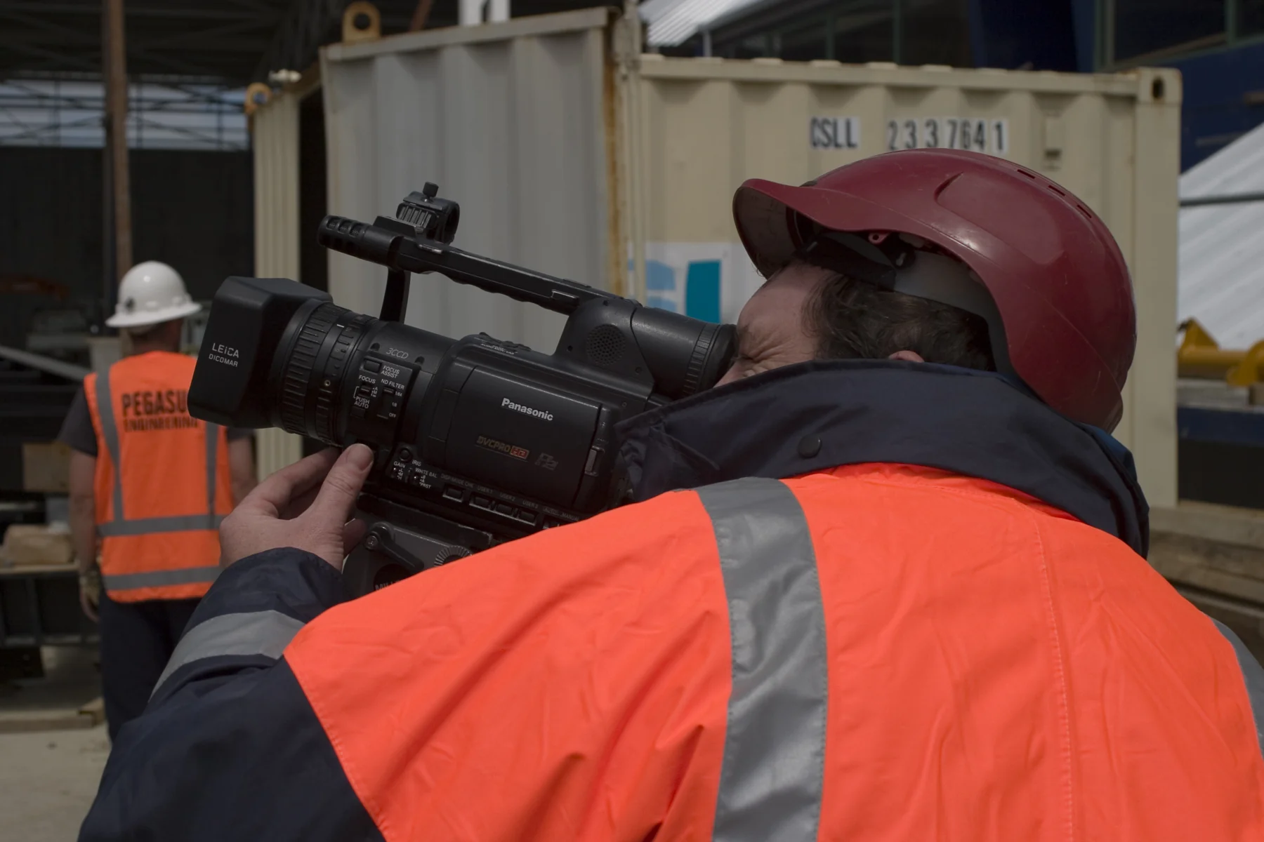 A camera operator in a hard hat and high-visibility jacket films activity at a construction site.