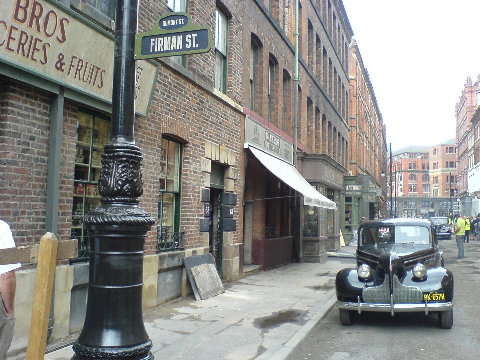 A Manchester street dressed as a period film set, with a vintage car, shop fronts and production staff.