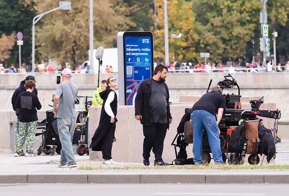 Camera and sound crew members make adjustments while filming on a riverside embankment.