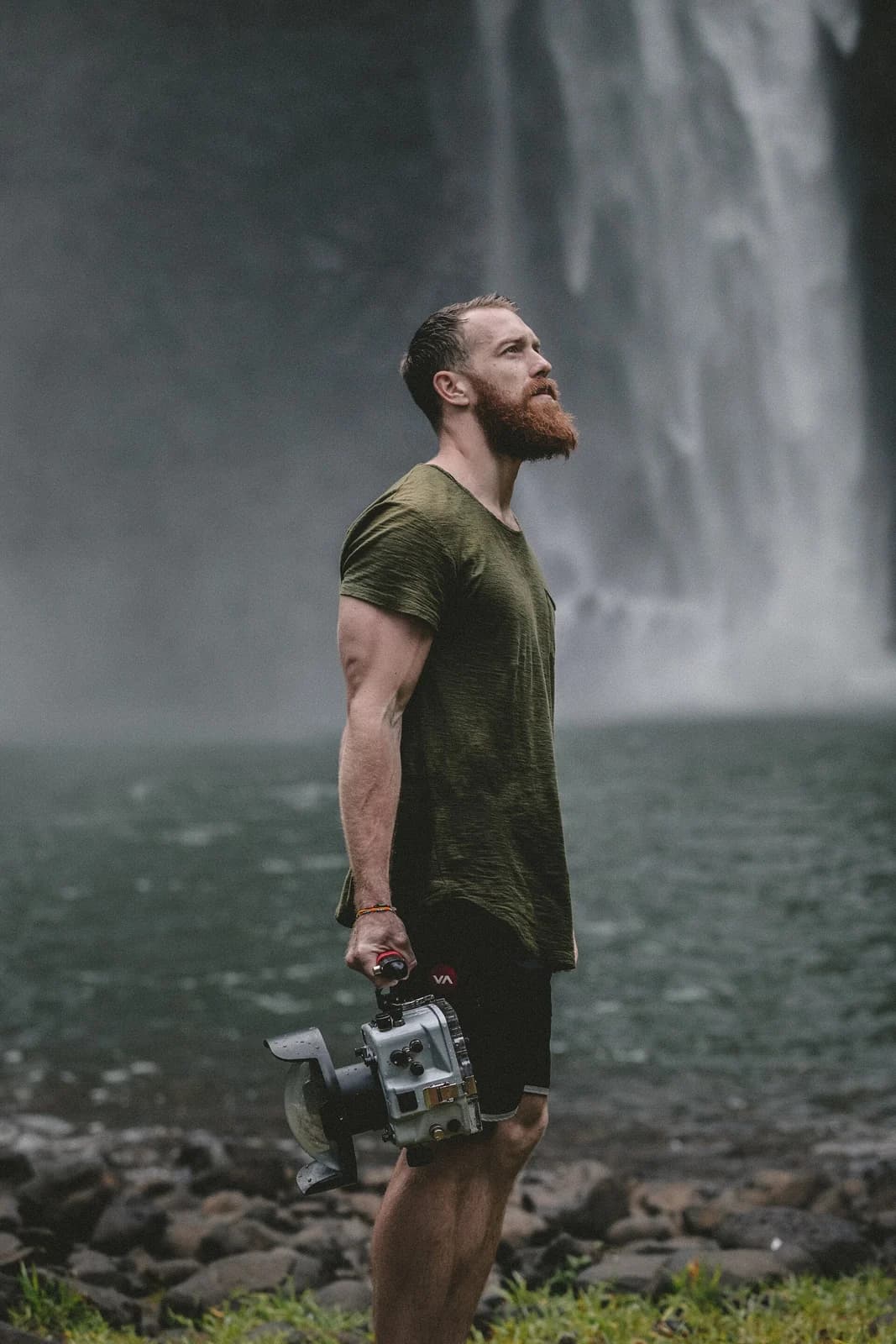 A filmmaker carries a camera in a rocky landscape beside a waterfall.