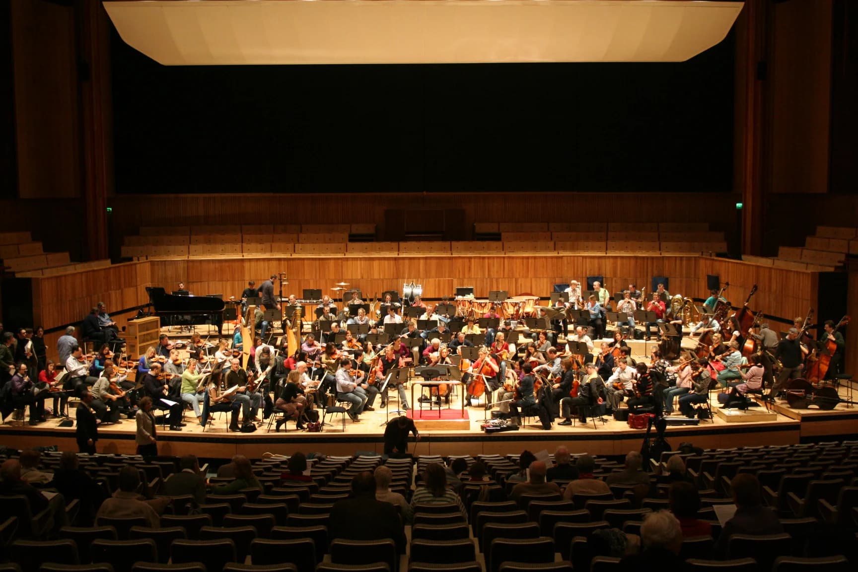 An orchestra rehearses on a concert stage with musicians, instruments and music stands.