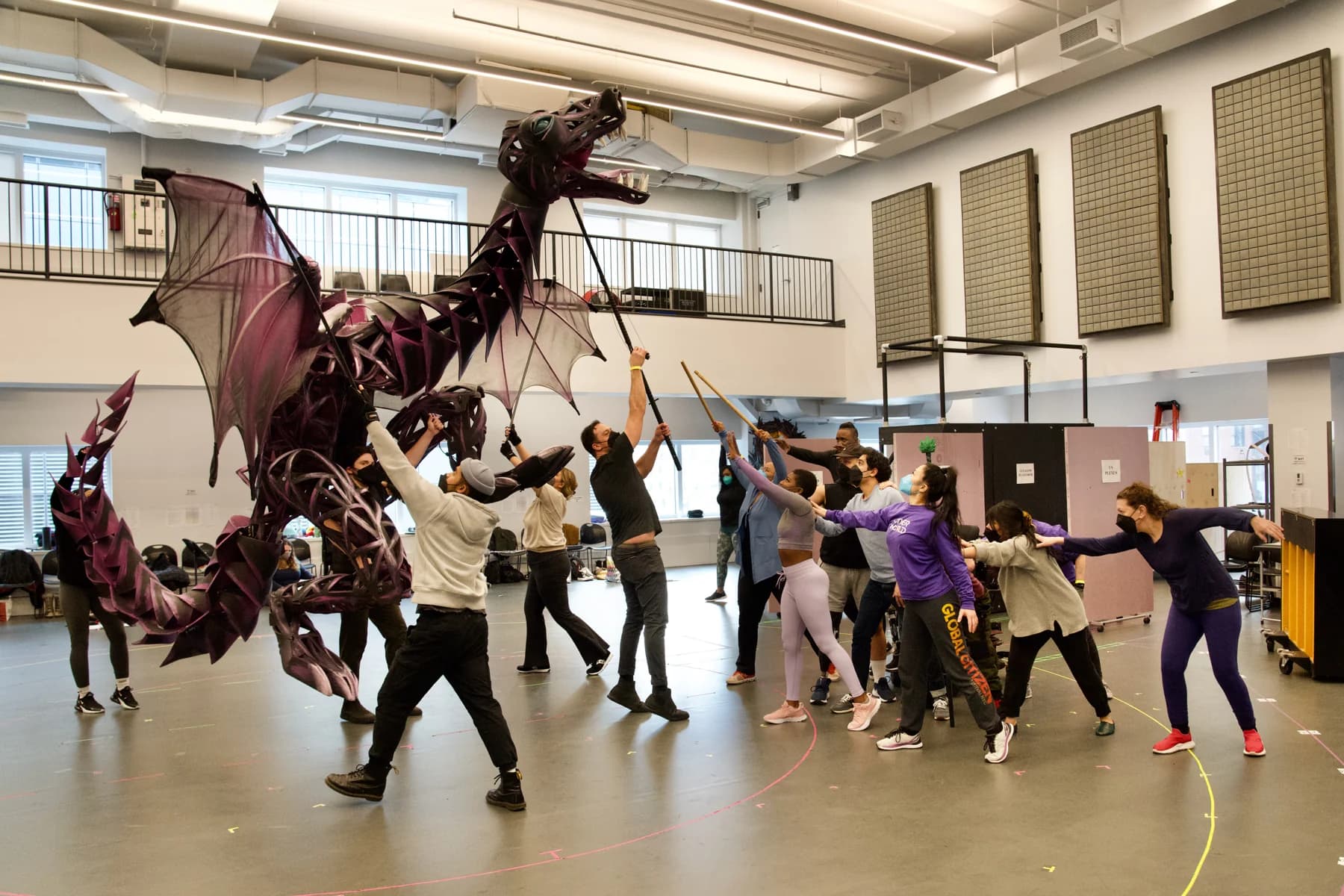 Performers rehearse together while operating a large theatrical dragon puppet in a rehearsal studio.