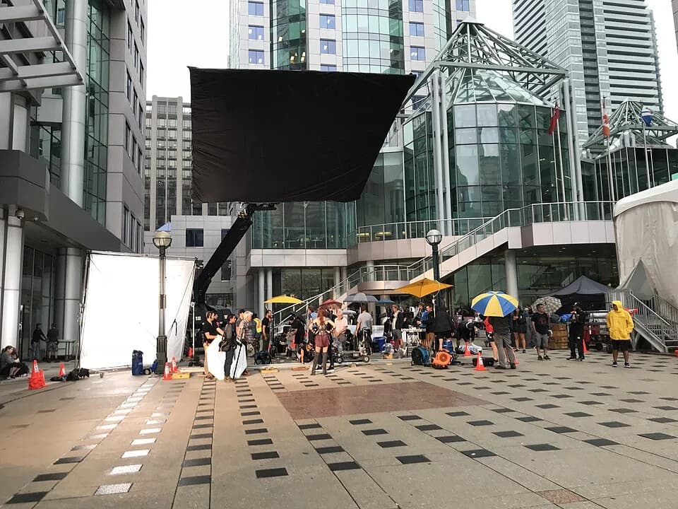 A film crew works on a city street beside a camera setup, overhead diffusion and lighting equipment.